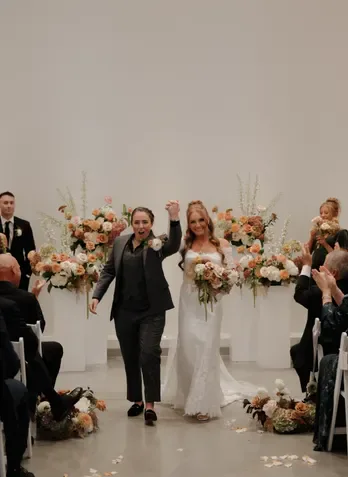 Two brides raising their fists in celebration as they walk back down a flower-lined aisle
